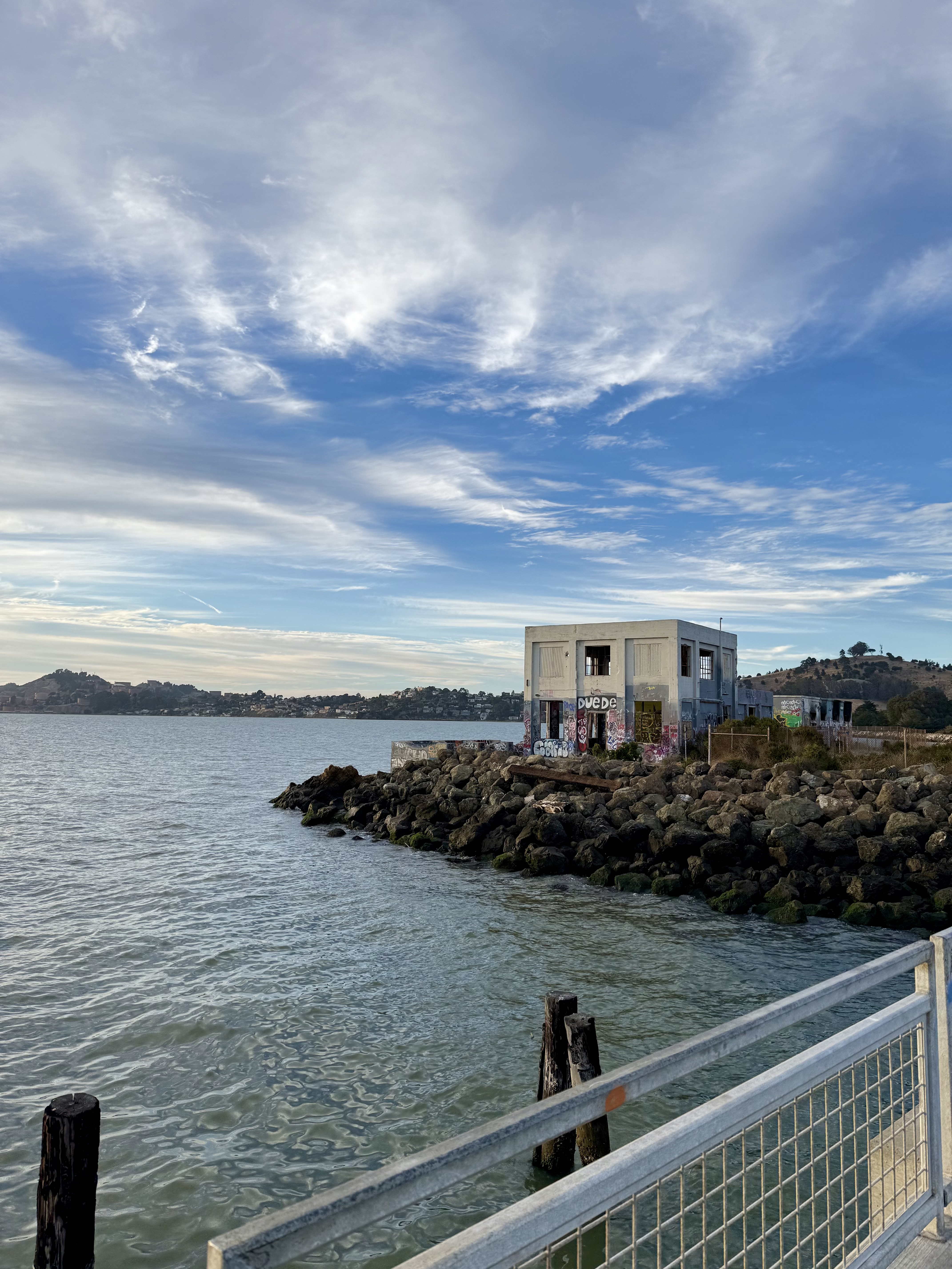 Calm waterfront with a graffiti-covered abandoned concrete building on a rocky shoreline, viewed from a pier with a metal railing, under a wide blue sky with wispy clouds.